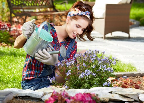 A woman with brown hair tied back and wearing a colorful plaid shirt, gardening gloves, and a headband, is kneeling on a landscaped garden bed within a residential outdoor space. She appears to be smiling and focused as she waters small flowering plants, likely lavender or similar, with a metal watering can. The garden features a curved stone border separating the flower bed from a neatly maintained lawn with dense, green grass. In the background, there are outdoor patio furniture pieces, including a wooden table and chairs, and a paved patio area, suggesting a well-kept backyard in London. Mature trees and shrubs are visible beyond the patio, adding natural privacy and greenery to the scene. The weather appears bright and sunny, with natural daylight illuminating the garden, which showcases a blend of flower beds, lush grass, and outdoor living space, reflecting typical features of garden maintenance and landscaping services provided by Great Gardeners in the London area within the postcode vicinity.