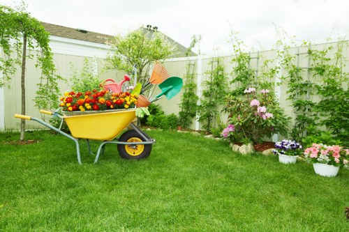 A view of a well-maintained backyard garden featuring a lush, evenly cut green lawn, bordered by a white fence adorned with green climbing plants and trailing vines. In the foreground, a yellow wheelbarrow filled with brightly coloured flowers and garden tools such as a watering can and small spade is visible, indicating recent gardening or maintenance activity. Along the right side, there are various flower beds with blooming plants in shades of pink, purple, and white, some planted directly in the soil while others are in individual white pots, creating vibrant colour contrasts against the greenery. A mature bush with pink flowers stands near the fence, contributing height and structure to the garden. The sky appears partly cloudy, suggesting mild weather, and the overall scene exudes a tidy, inviting outdoor space, suitable for lawn care and gardening services provided by companies like Great Gardeners in London.