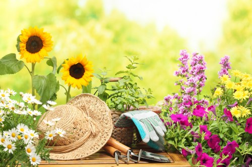 A vibrant outdoor garden scene featuring a variety of flowering plants and gardening tools. In the foreground, there are bright yellow sunflowers with dark centers, alongside clusters of white daisies with yellow centers and pink petunias, all adding natural colour to the space. A wooden table, partially visible, holds gardening accessories including a straw hat, garden gloves, and small hand tools such as pruning shears and a trowel, suggesting recent gardening activity. The background showcases a lush, blurred green garden with shrubs and trees, indicating a well-maintained outdoor area typical of a UK suburban or residential garden near London, with a bright and pleasant weather condition, possibly in late spring or summer. The scene exudes a healthy and thriving garden environment, suitable for lawn care, planting, and ongoing outdoor maintenance by professionals like Great Gardeners based in London, highlighting elements relevant to local gardening services like lawn mowing, plant care, and seasonal planting in a landscaped setting.